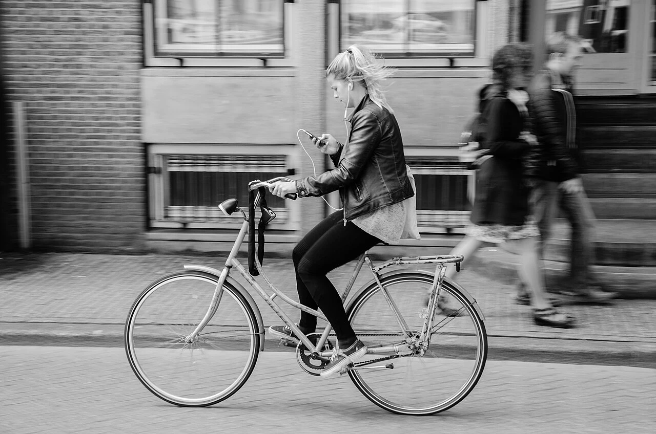 Black and white photo of a woman riding a bicycle on a city street with earbuds in, looking at her phone