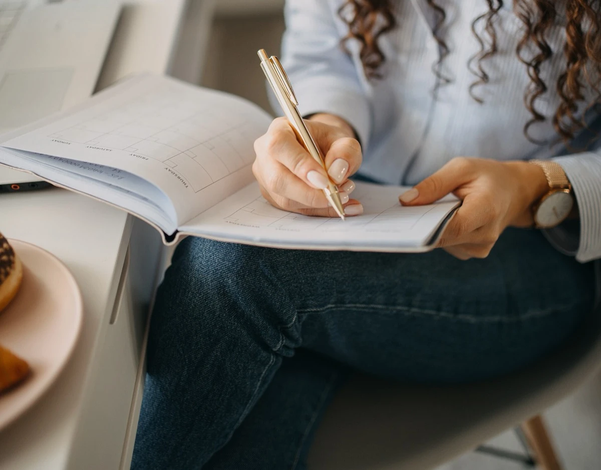 A woman writes notes with a gold pen in a planner open on her lap